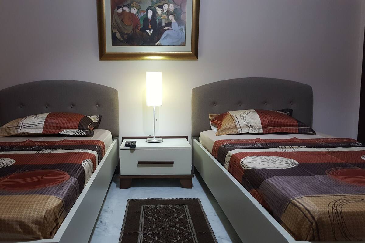 Ein Hotelzimmer mit zwei Betten, jeweils mit einem gemusterten Überwurf, einem Nachttisch mit Lampe und einem Gemälde an der Wand.