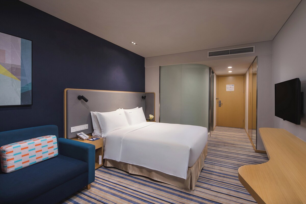 Quarto de hotel com cama, escrivaninha, cadeira, televisão e um quadro na parede.