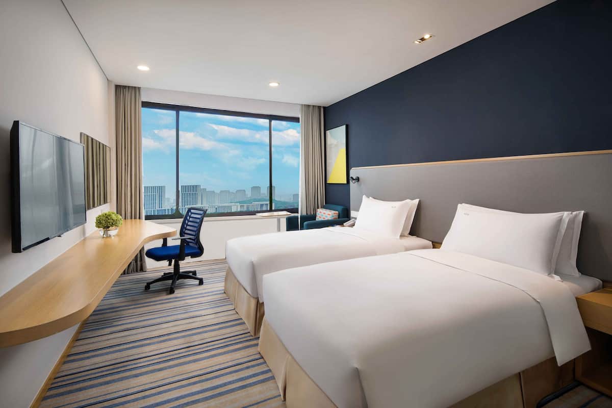 Ein Hotelzimmer mit zwei Betten, einem Schreibtisch, einem Sessel, einem Fernseher und einem großen Fenster mit Stadtblick.