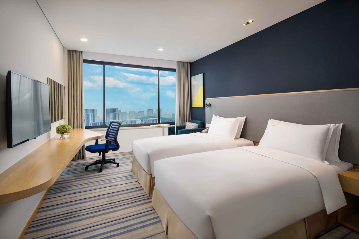 Quarto de hotel com duas camas, uma escrivaninha, uma cadeira, televisão e uma janela grande com vista para a cidade.
