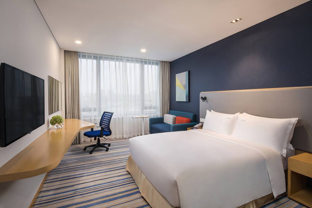 Ein Hotelzimmer mit einem großen Bett, einem Schreibtisch mit Stuhl, einem Fernseher und einem Fenster mit Vorhängen.