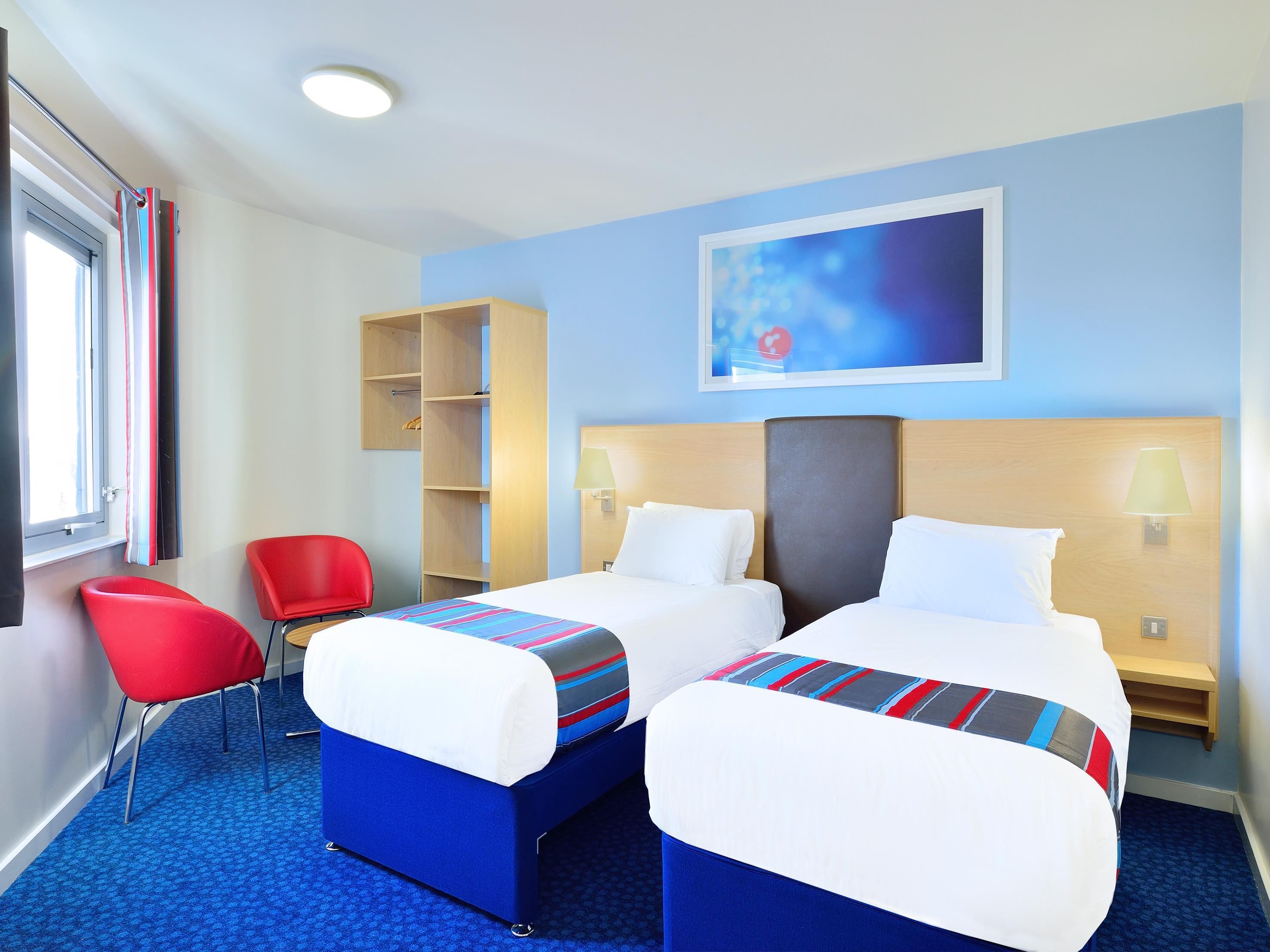 A hotel room with two beds, a red chair, a wooden shelf, and a painting on the wall.