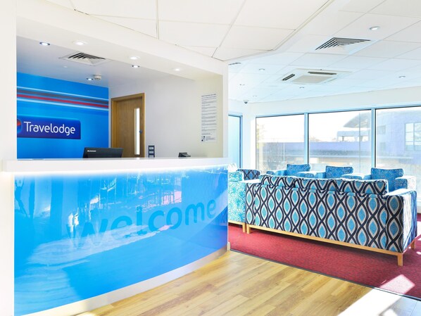A reception area with a 'Travelodge' sign, a reception desk, a seating area with patterned cushions, and large windows offering a view. - Travelodge Birmingham Maypole (Birmingham)