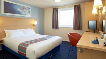 A hotel room with a bed, a desk with a lamp, a television, and a window with curtains.