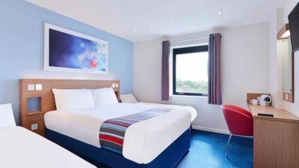 Una camera d'albergo con un letto, una scrivania, una sedia, una finestra con tende e un quadro appeso al muro.