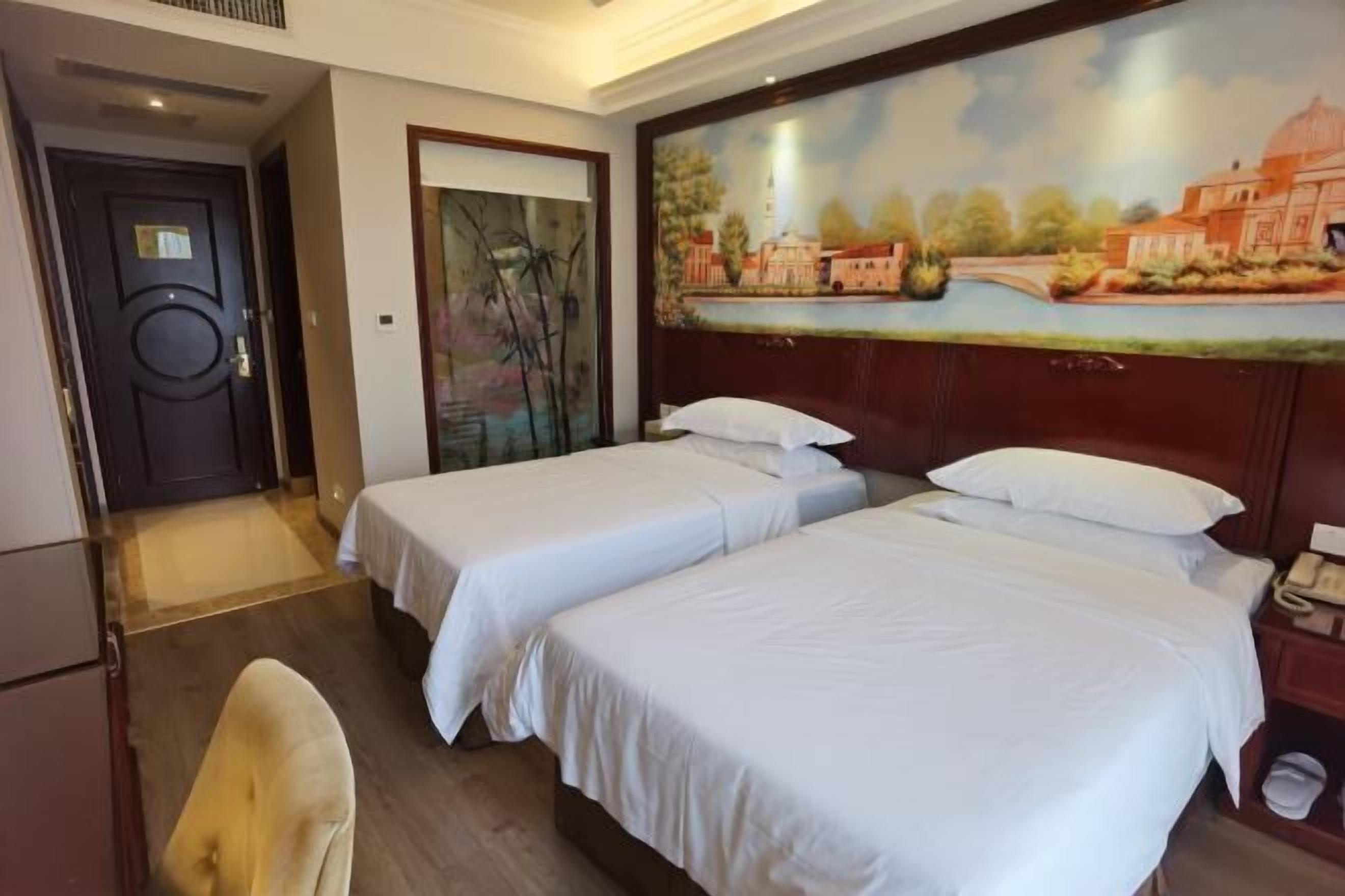 Une chambre d’hôtel avec deux lits simples, une tête de lit en bois, un tableau représentant un paysage, une porte et une chaise.