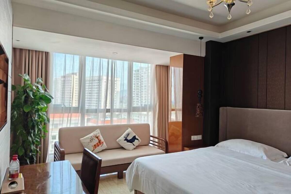 A hotel room with a bed, a sofa with cushions, a dining table, and a potted plant.