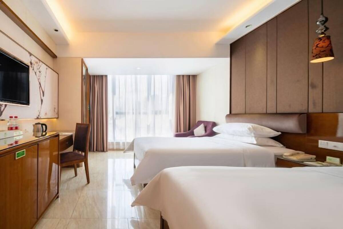 A hotel room with two beds, a desk, a chair, a TV, and a window with curtains.