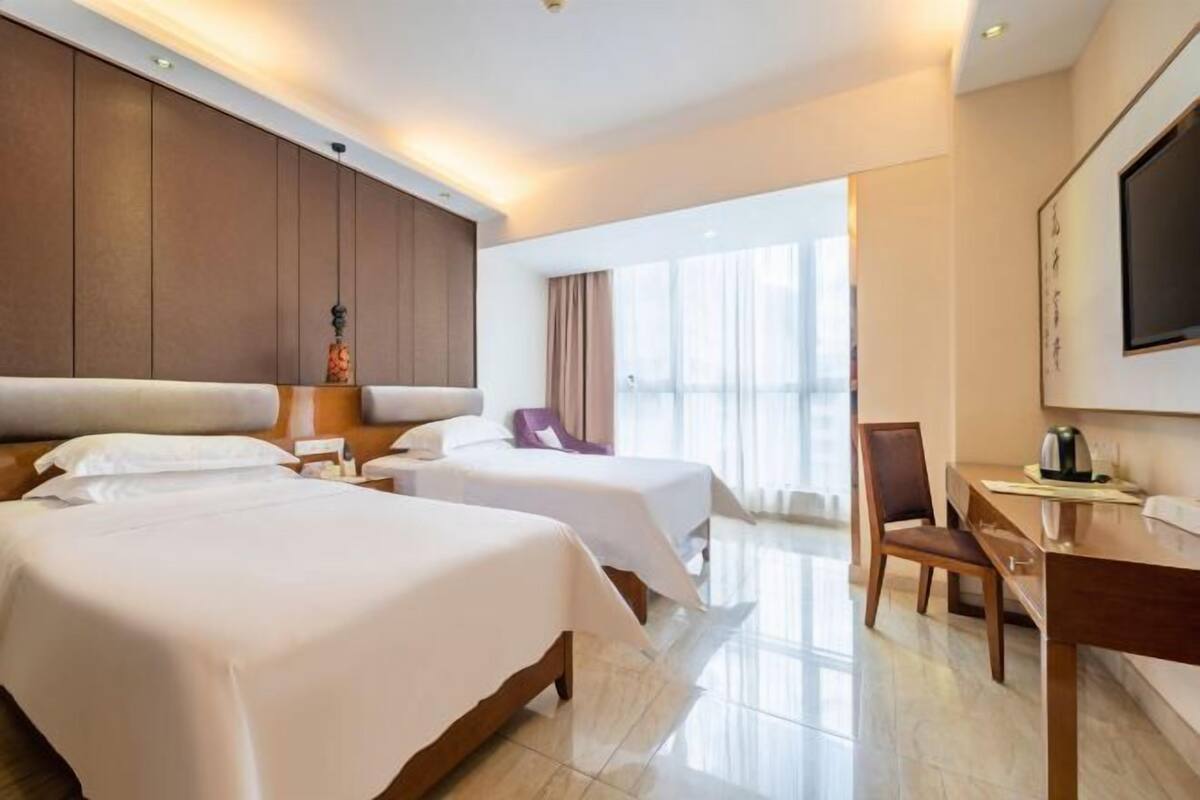 A hotel room with two beds, a desk, a chair, a TV, and a large window.