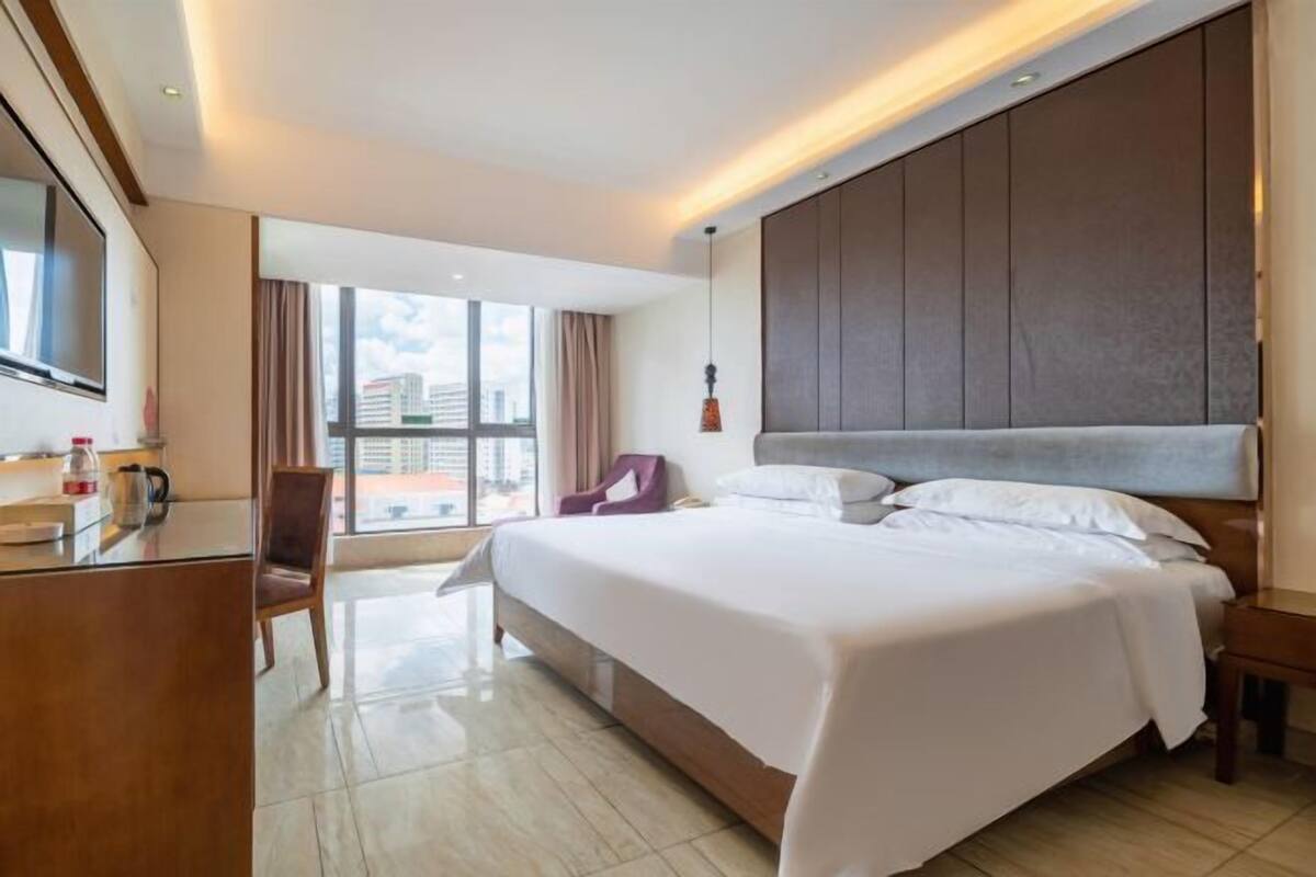 A hotel room with a large bed, a desk with a chair, a TV, and a window with a city view.