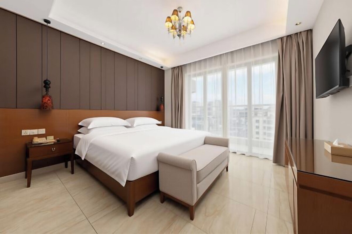 A hotel room with a large bed, a bedside table, a TV, and a view of the city.