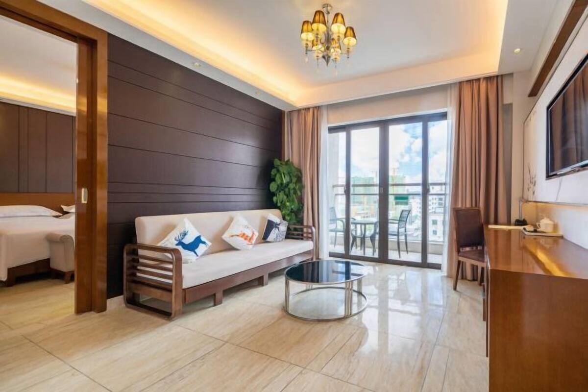 A modern hotel room with a sofa, a small round table, a balcony with a view, and a TV on the wall.