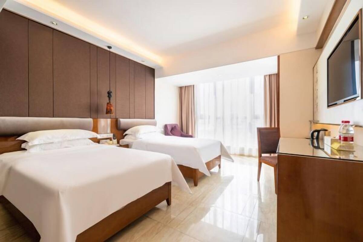 A hotel room with two beds, a desk, a TV, and a large window.