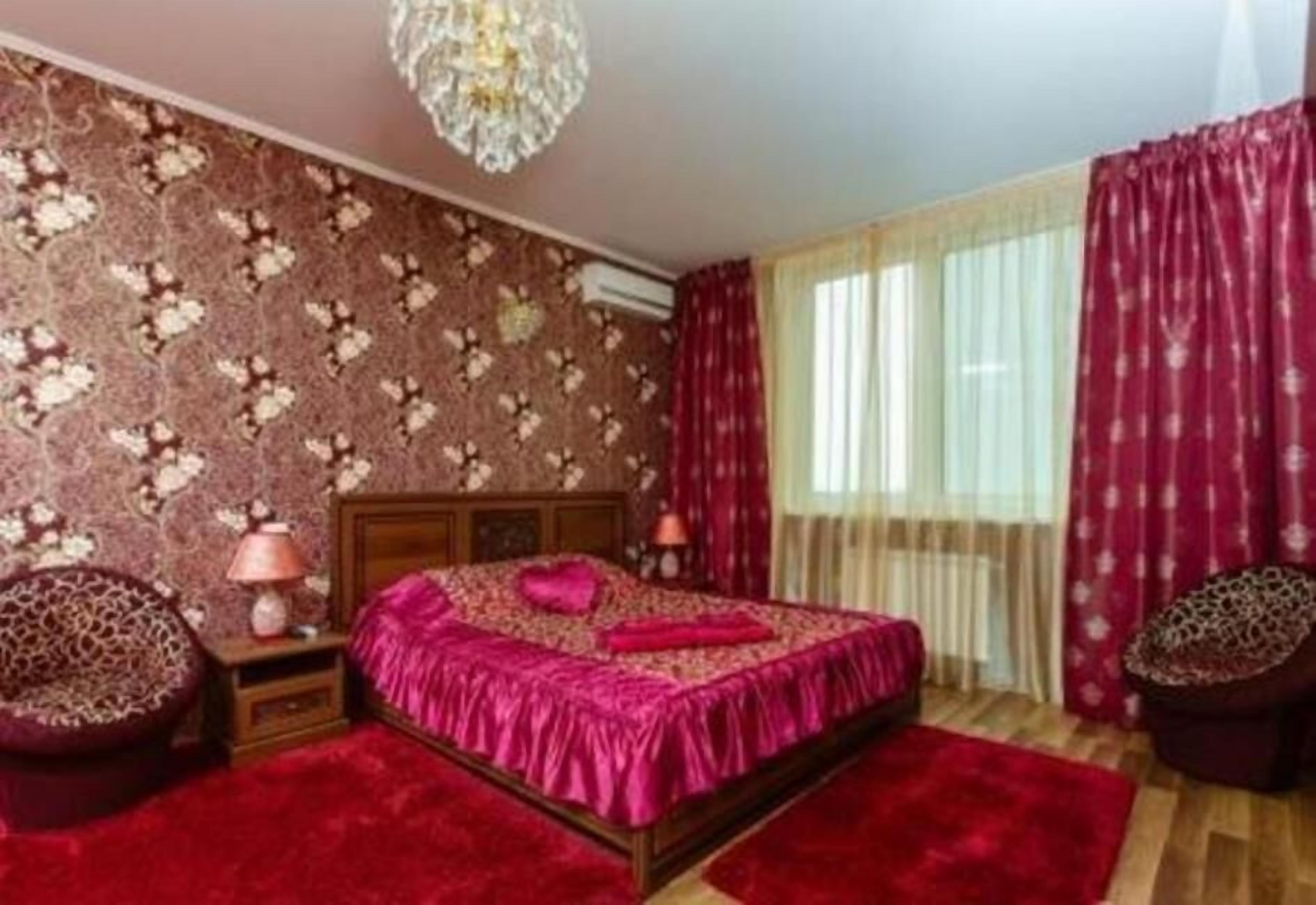 A bedroom with a floral wallpaper, a red bedspread, a chandelier, and a red rug.
