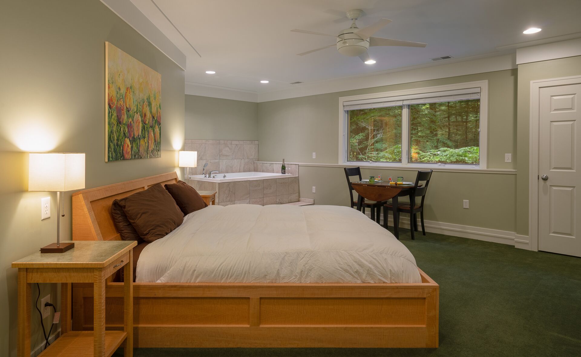 A bedroom with a bed, bedside table, lamp, painting, bathroom, window, and a view of trees.