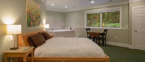 A bedroom with a bed, bedside table, lamp, painting, bathroom, window, and a view of trees.