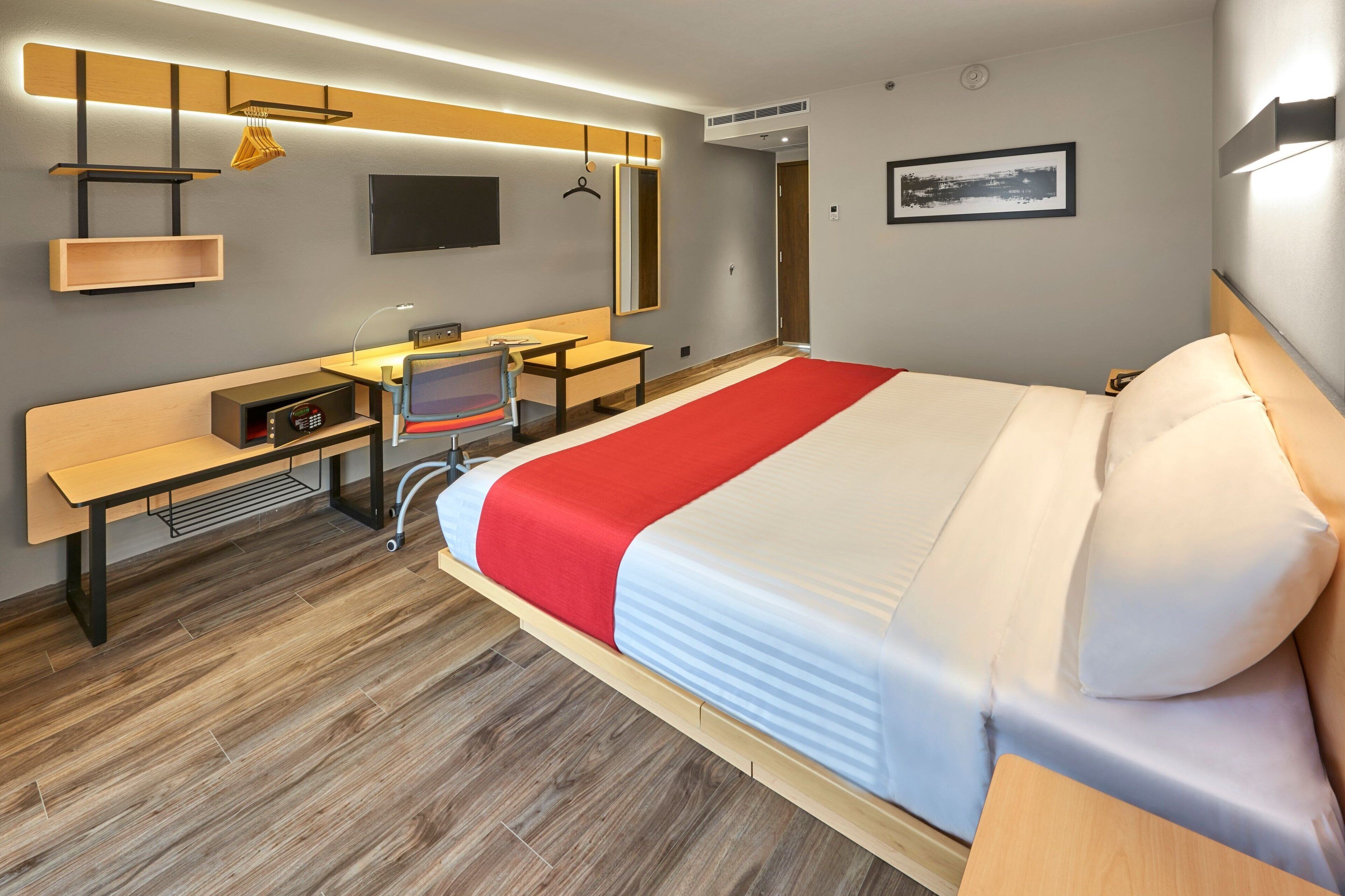 A modern hotel room with a large bed, a desk, a chair, a television, and a wardrobe.
