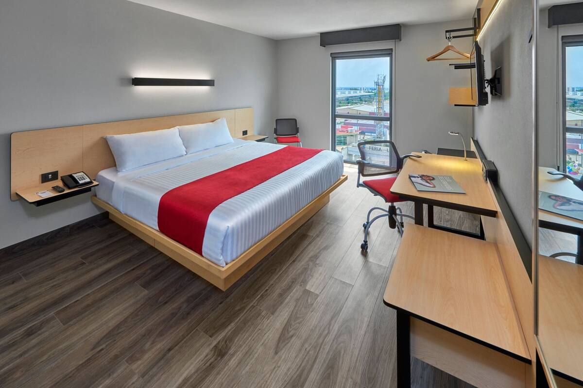 A modern hotel room with a large bed, a desk, and a chair. There is a window with a view of buildings and a cityscape.