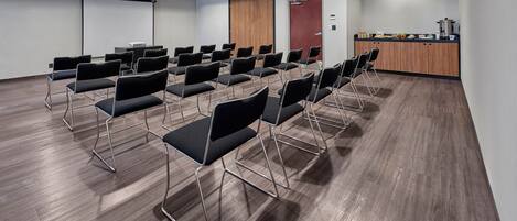 A conference room with rows of black chairs, a projector screen, a wooden door, and a kitchenette area.