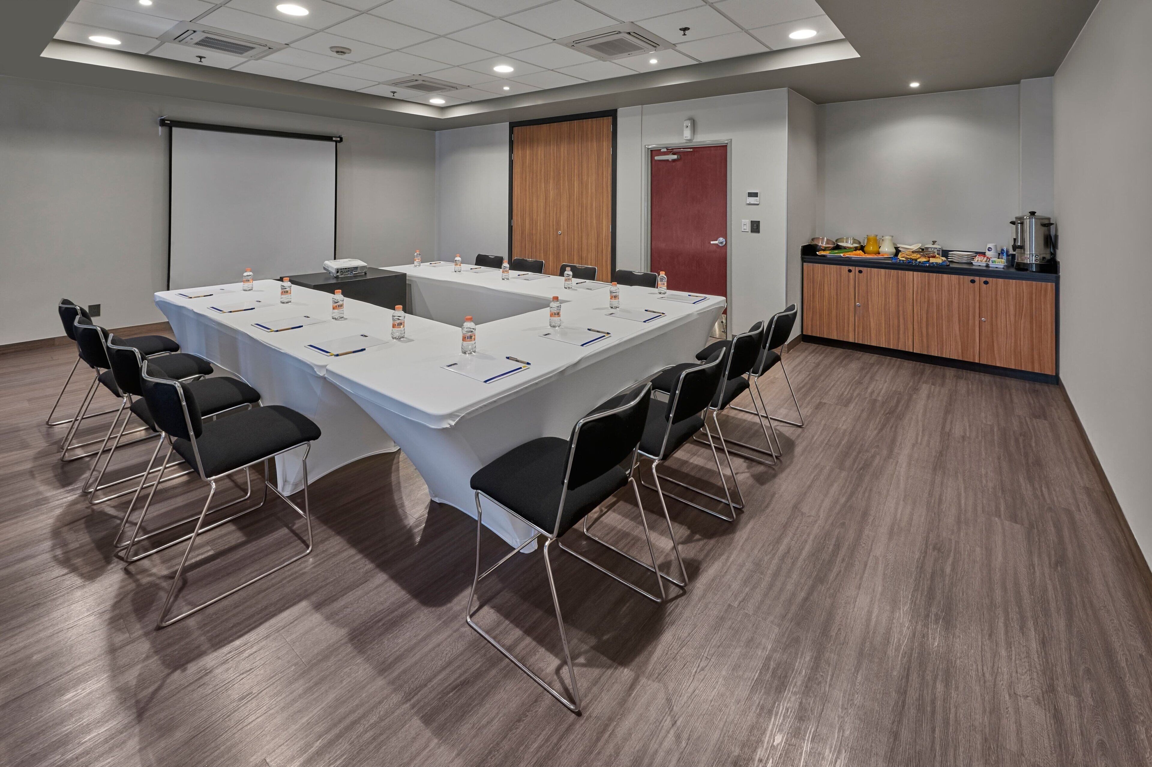Une salle de conférence avec une longue table, des chaises noires, un écran de projection, une porte en bois et un coin cuisine.