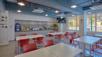 Eine moderne Cafeteria mit weißen Tischen und roten Stühlen, einem Küchenbereich und großen Fenstern mit Blick nach außen.