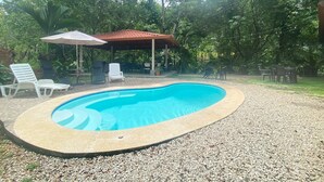 Une piscine dotée d’un espace détente couvert, entourée d’arbres et de gravier.