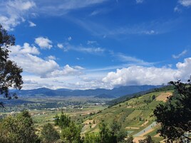 A panoramic view of a hilly landscape with a river, green fields, and distant mountains under a clear blue sky.