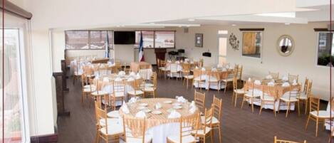 A spacious banquet hall with round tables and chairs, a stage with a podium, and a large window providing natural light.