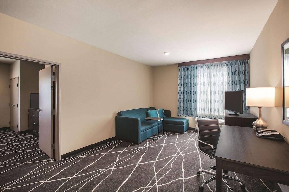 A hotel room with a sofa, a desk with a lamp, a TV, and a patterned carpet.