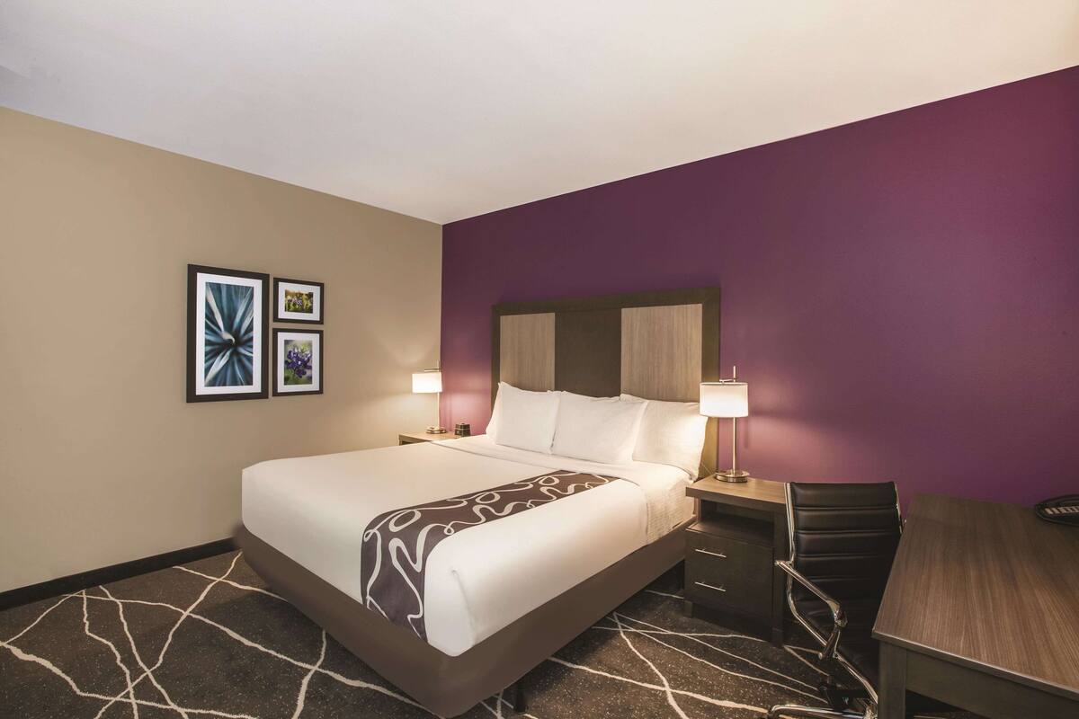 A hotel room with a large bed, two bedside tables, a desk, and a chair. There are two wall-mounted lamps and a framed picture on the wall.