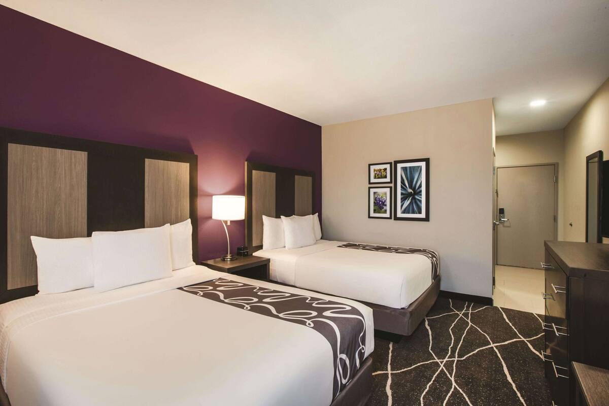 A hotel room with two beds, a nightstand with a lamp, a dresser, and framed artwork on the wall.