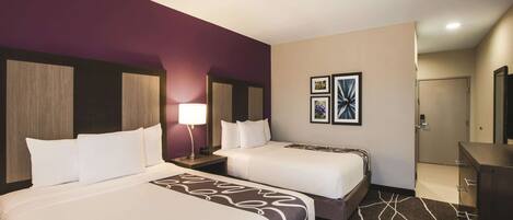 A hotel room with two beds, a nightstand with a lamp, a dresser, and a door leading to another room.
