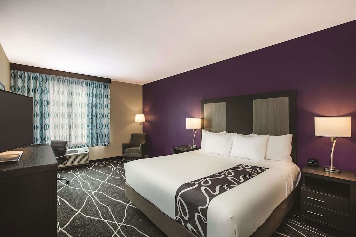A hotel room with a large bed, two bedside lamps, a desk with a chair, a TV, and a window with patterned curtains.