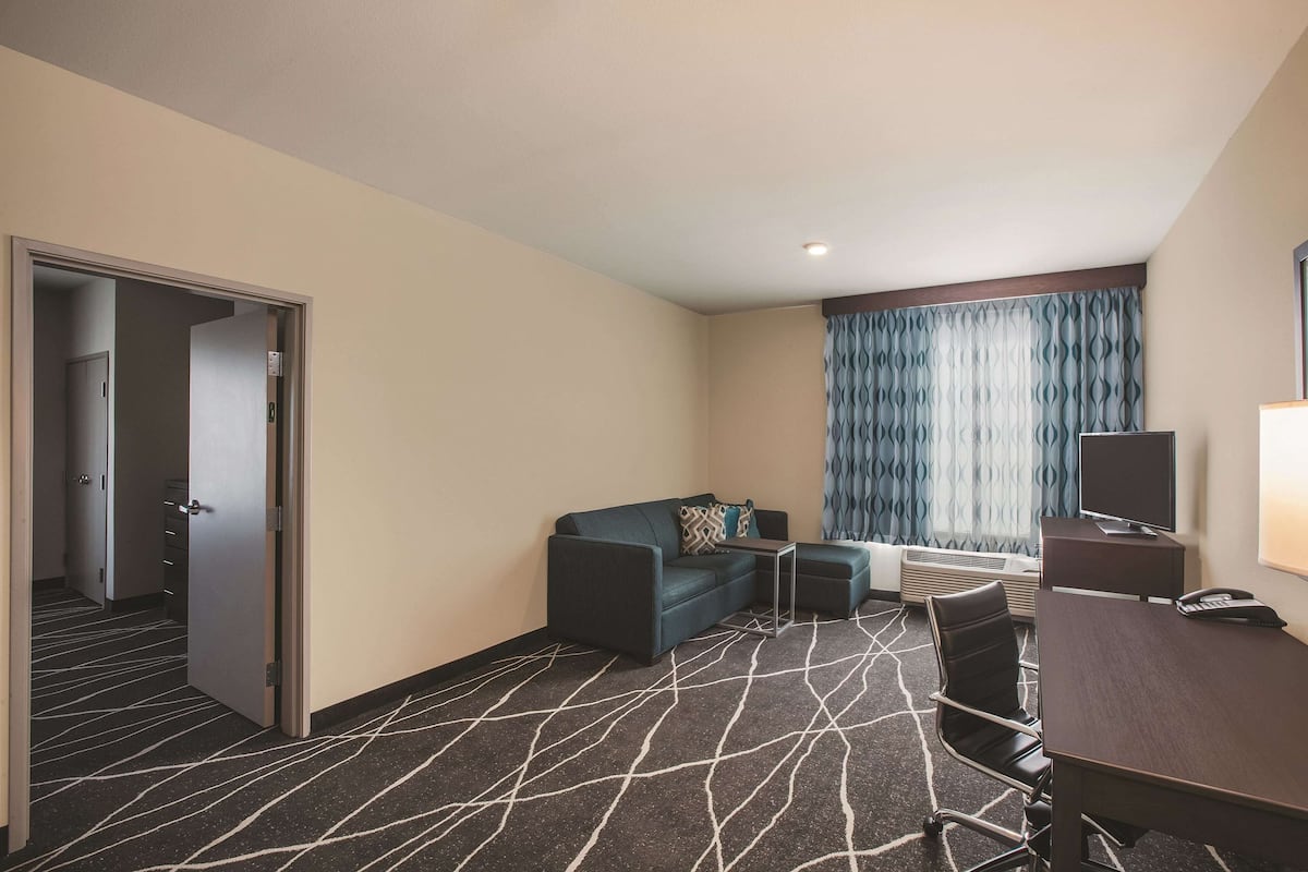 A hotel room with a dark patterned carpet, a sofa, a small table, a desk, a chair, a TV, and a window with curtains.