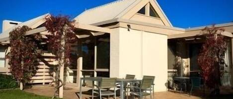 Une maison moderne avec un toit en pente, de grandes fenêtres et une terrasse aménagée avec du mobilier d’extérieur.