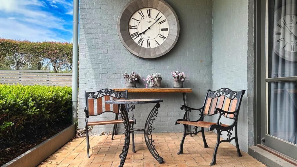 A patio with a round wall clock, two chairs, a small table, and a shelf with flowers.