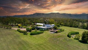 A large, modern house with solar panels, surrounded by well-maintained lawns and gardens, set against a backdrop of mountains and a sunset sky. - Grapevines Boutique Accommodation (Pokolbin)