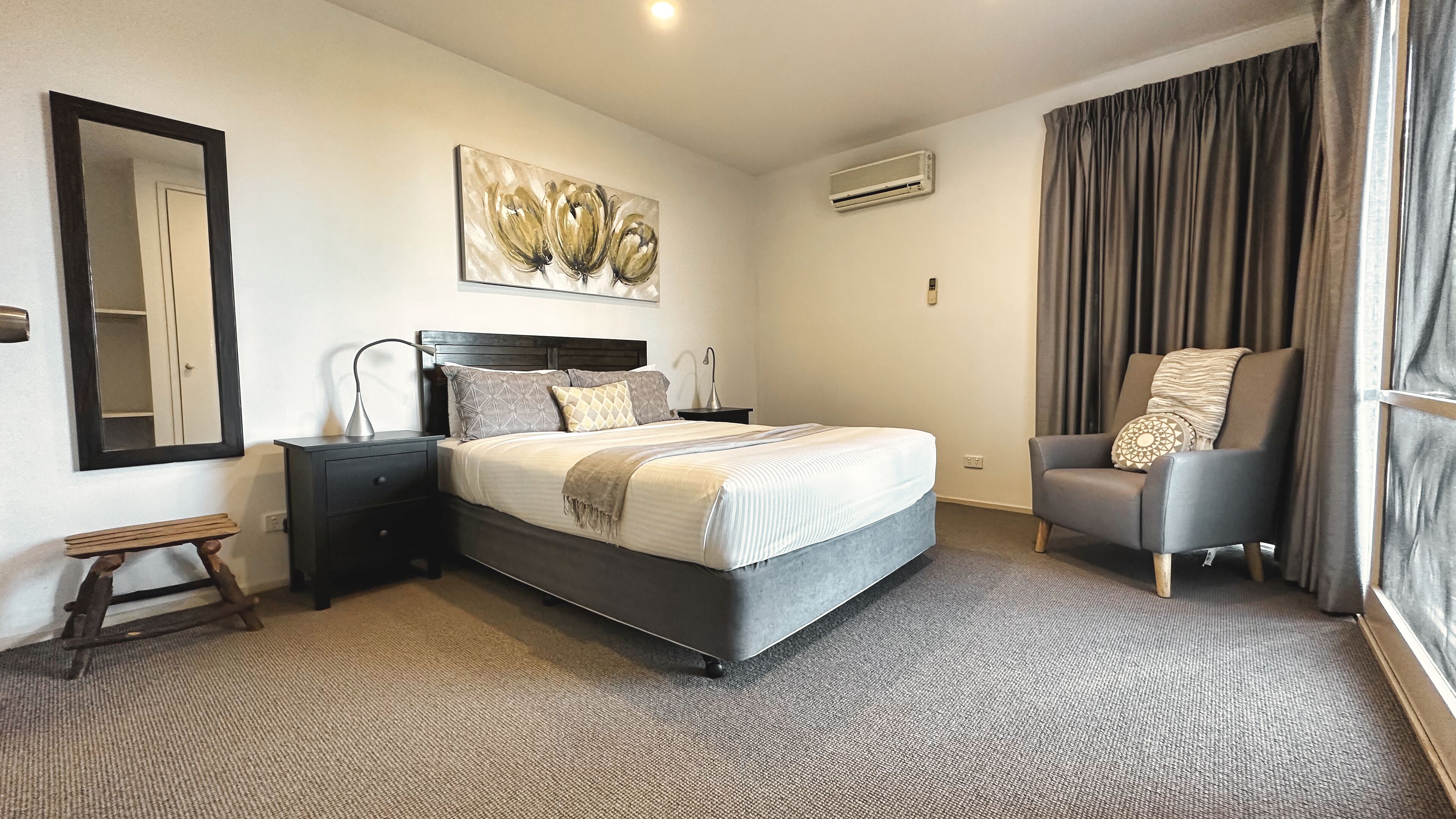 Habitación de hotel con una cama grande, una mesita de noche con lámpara, una silla, un espejo y un cuadro en la pared.