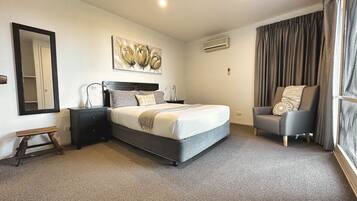 Habitación de hotel con una cama grande, una mesita de noche con lámpara, una silla, un espejo y un cuadro en la pared.