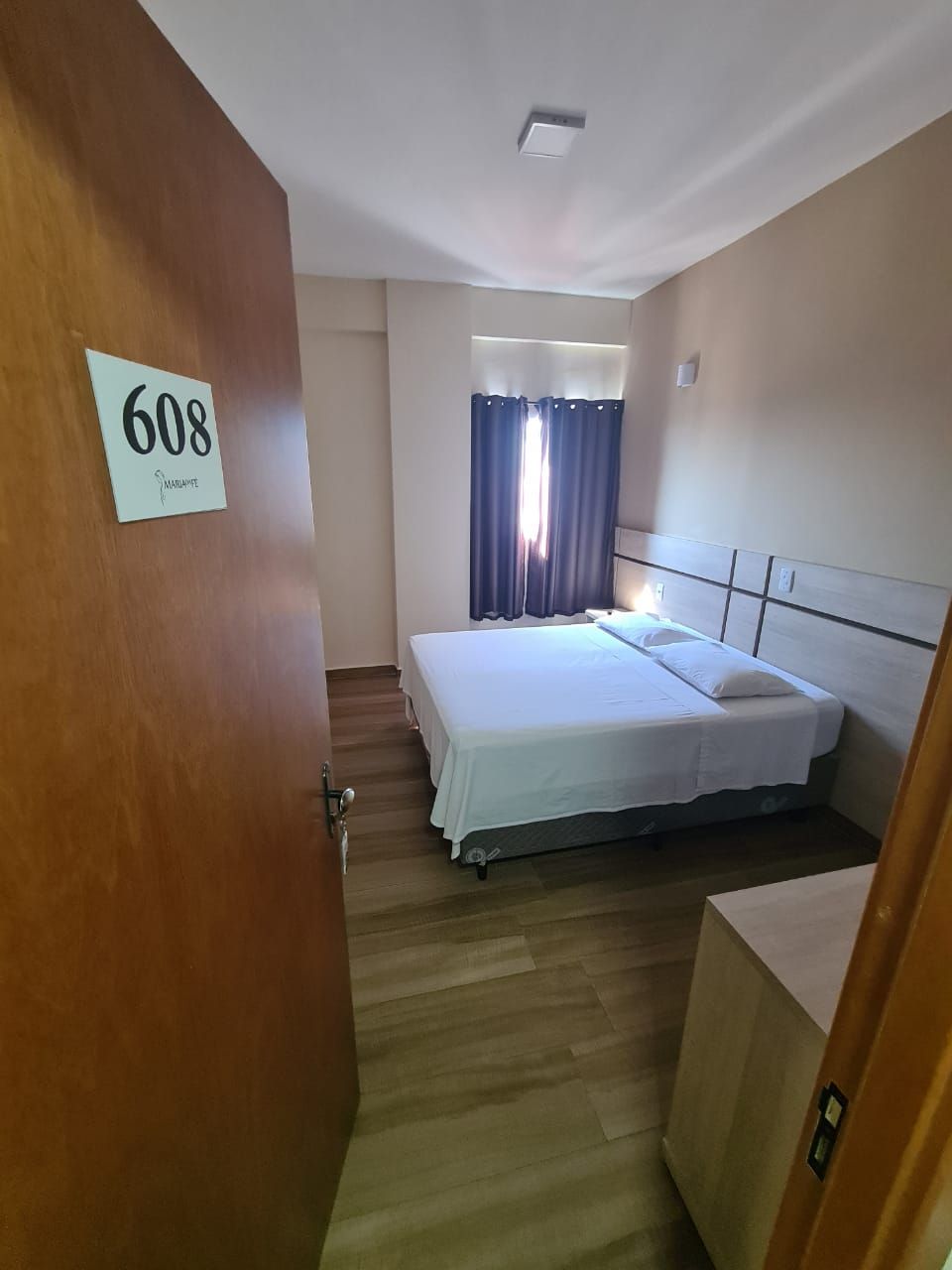 Egy szállodai szoba, amelyben található egy ágy, egy éjjeliszemély, egy számjelzéssel ellátott fa ajtó és egy függönnyel ellátott ablak.