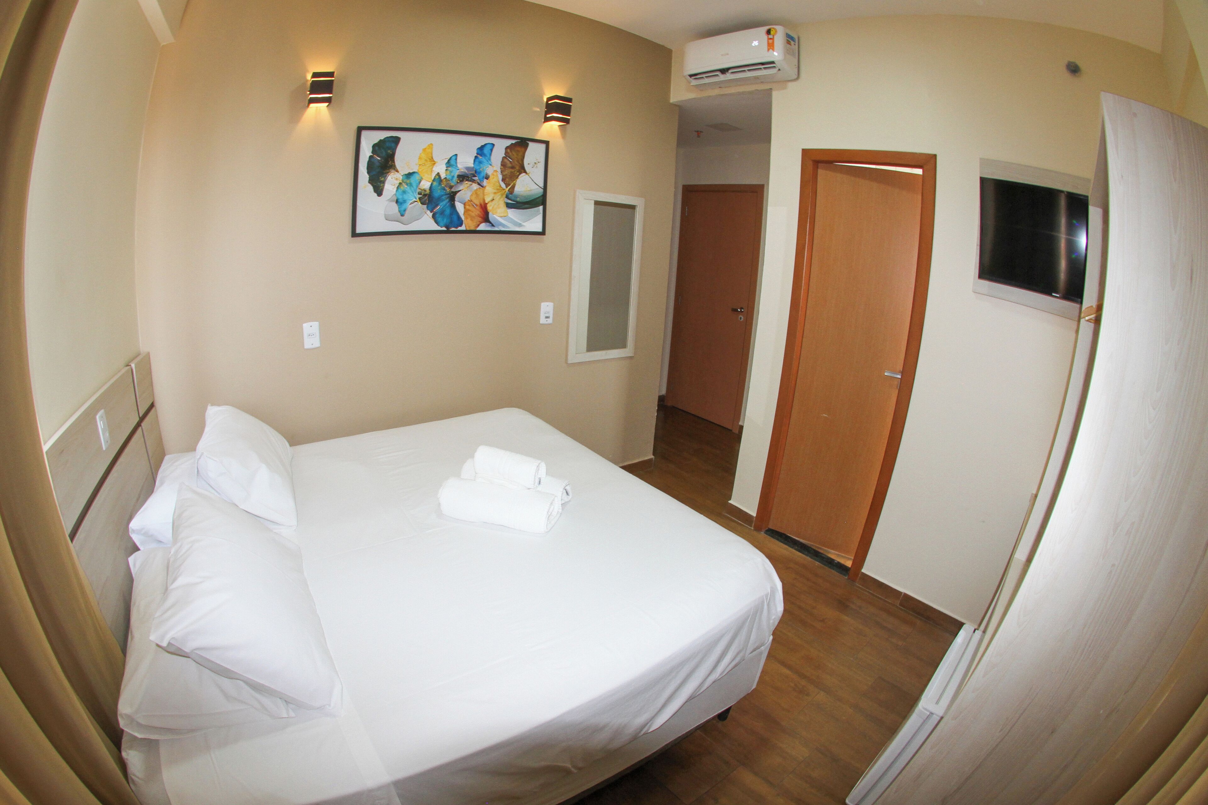A hotel room with a bed, a painting on the wall, an air conditioning unit, and a door leading to another room.