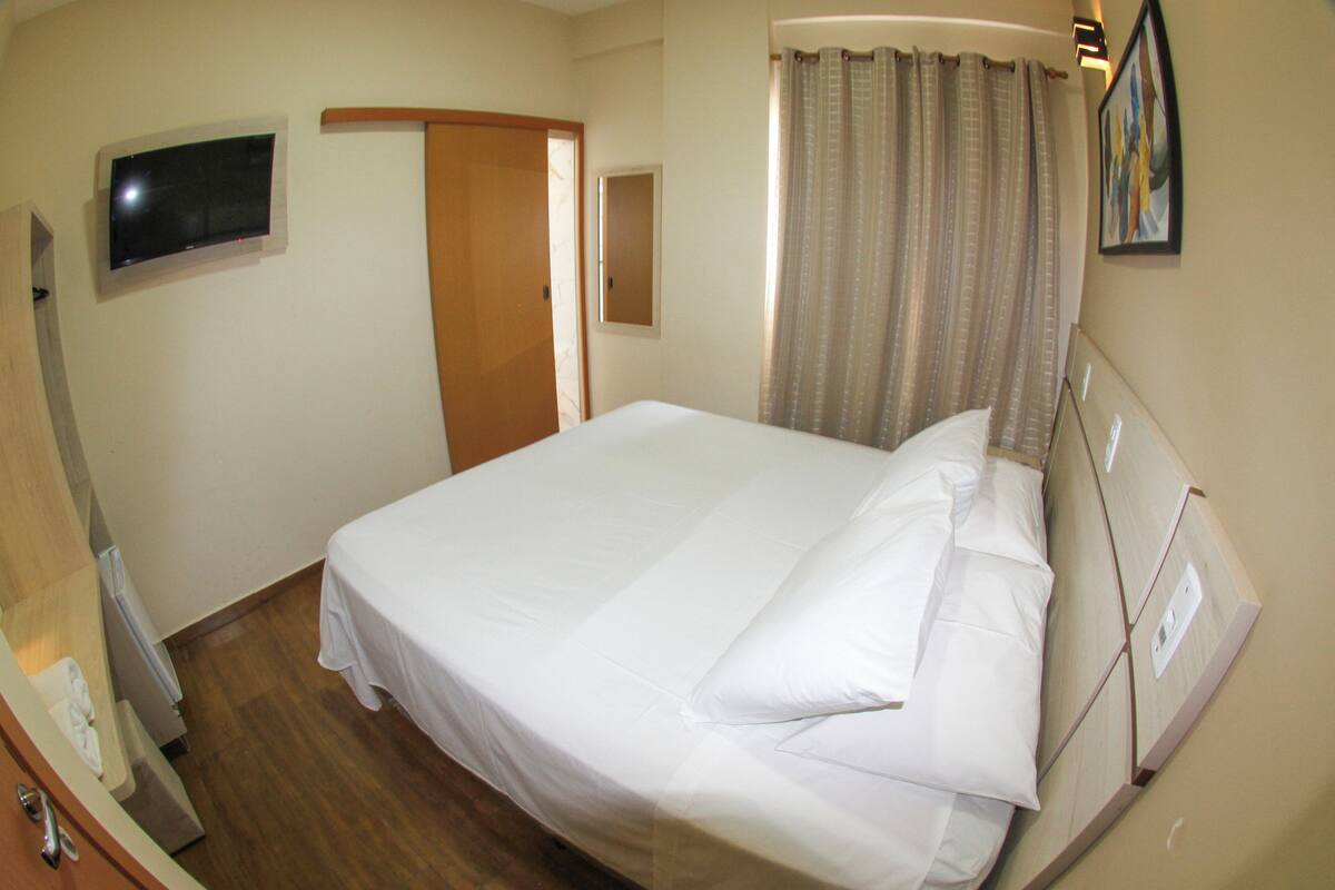Una habitación de hotel con una cama, un televisor fijado a la pared, una puerta y una ventana con cortinas.