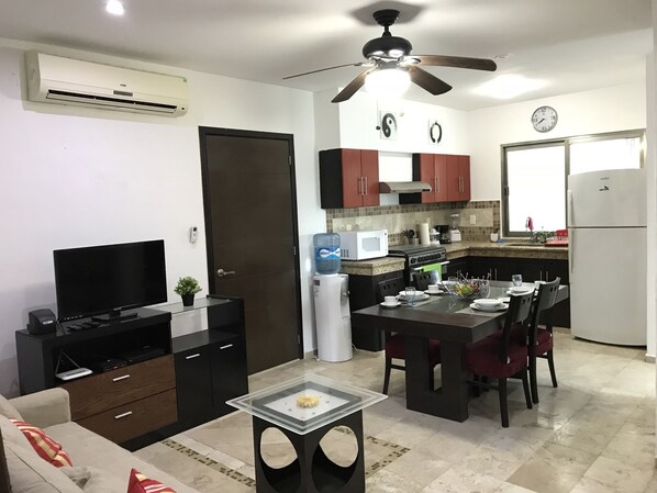 A modern kitchen with a microwave, stove, and refrigerator. There is a dining table with chairs and a small table with a decorative item. A ceiling fan is visible above the kitchen. - Lilas del Mar (Playa del Carmen)
