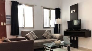 Una sala de estar con sofá, sillón, mesa de centro de vidrio y televisor sobre un mueble.