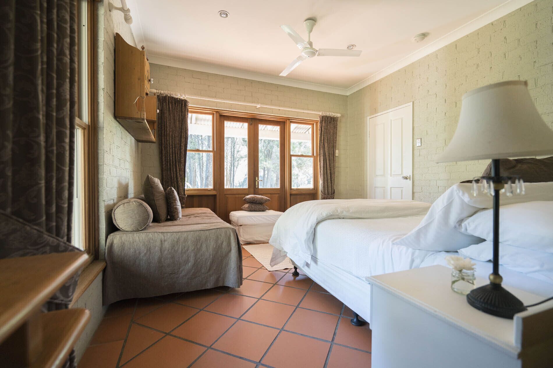 Un dormitorio con cama, una mesita de noche con lámpara, ventana con cortinas, puerta y ventilador de techo.