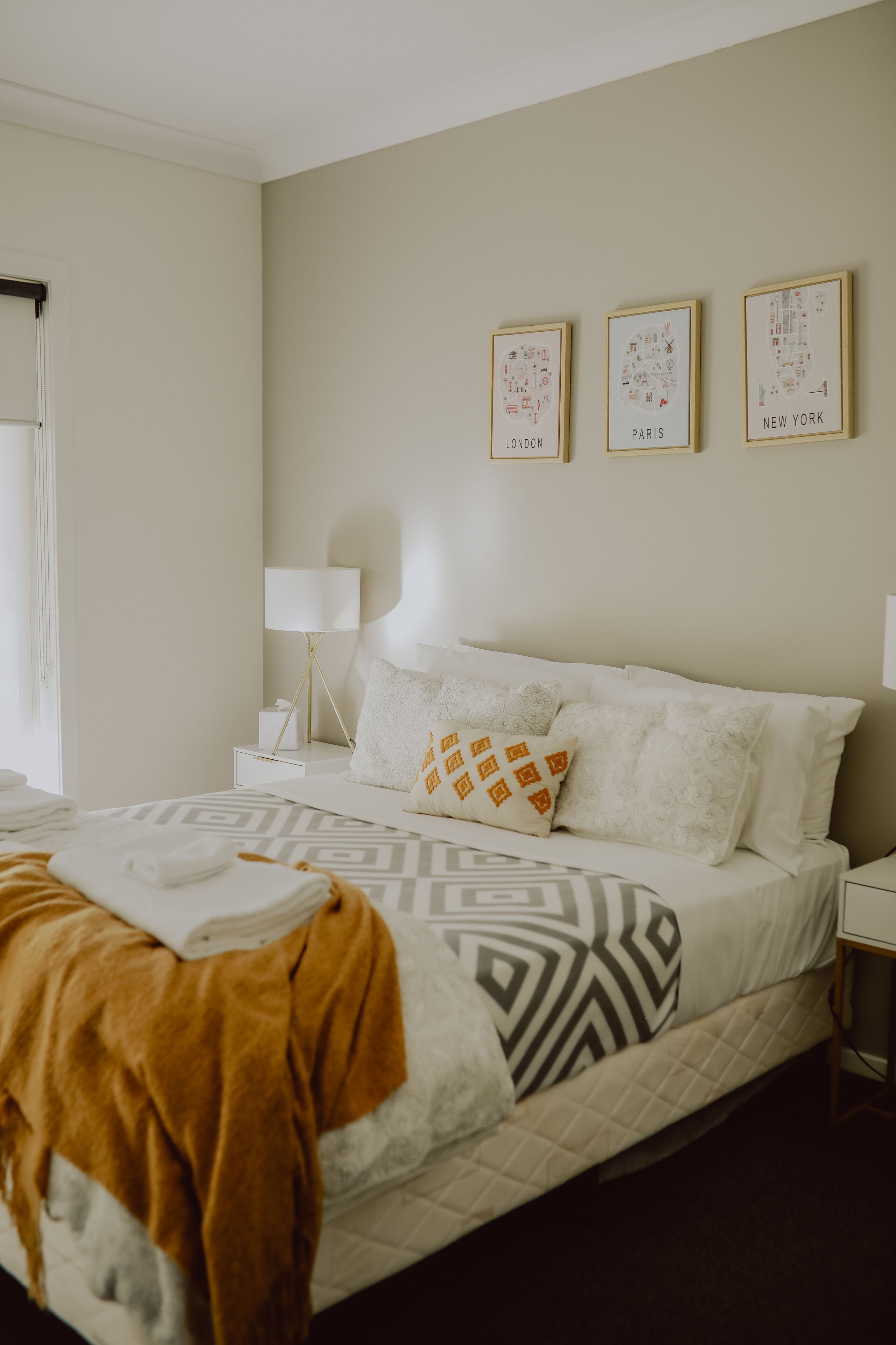 A neatly arranged hotel room with a bed, a bedside table with a lamp, and three framed city maps on the wall.