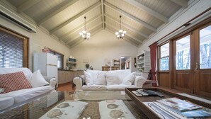 A cozy living room with a white sofa, a glass coffee table, and a wicker side table with magazines. A kitchen with wooden cabinets and a microwave is visible in the background. - Thistle Hill Guest House (Pokolbin)
