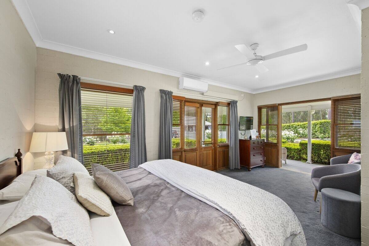 A bedroom with a large bed, a desk, and a chair. There are windows with curtains and a view of greenery outside.