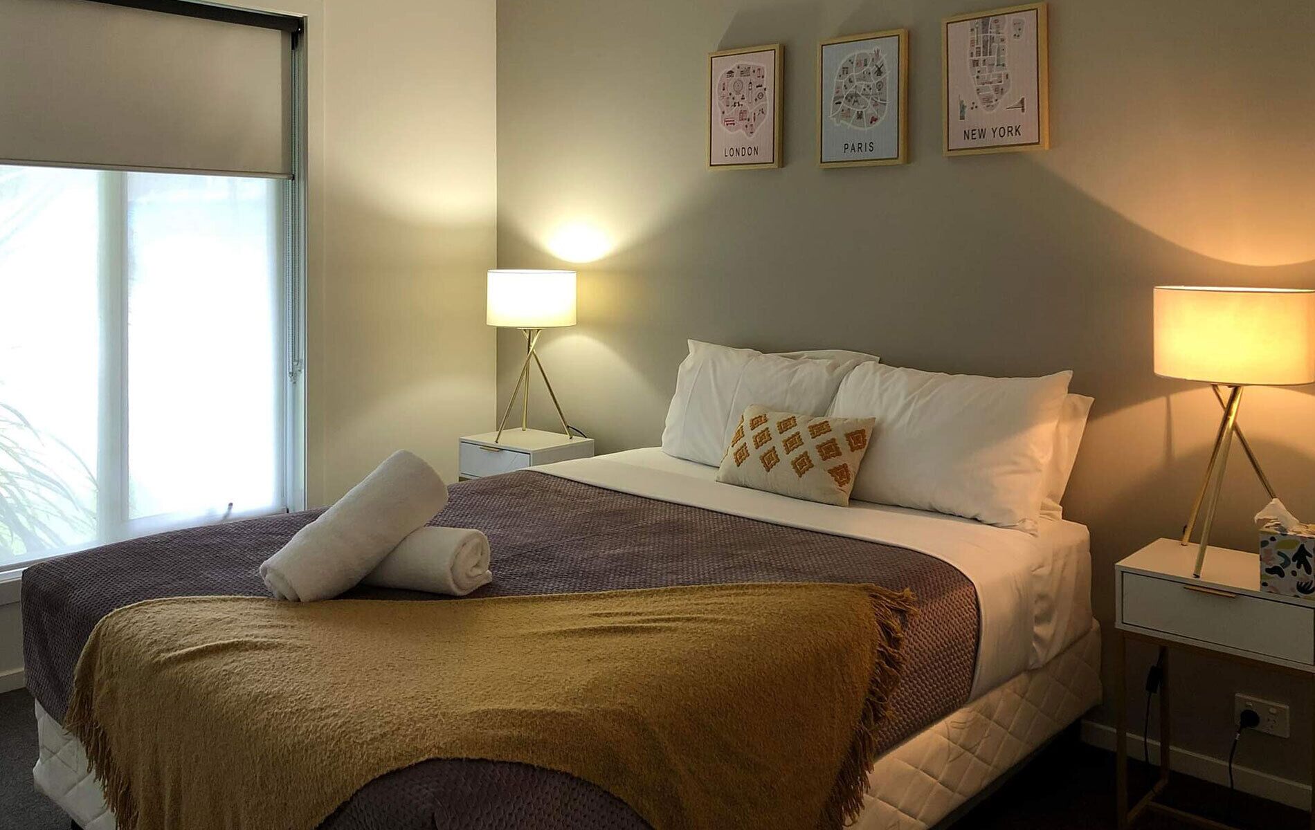A hotel room with a bed, two bedside tables, a window, and wall art depicting London, Paris, and New York.