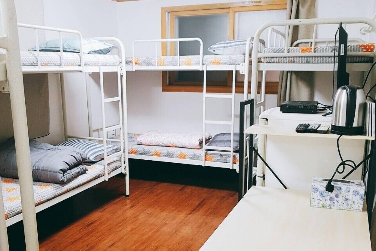 A dormitory room with bunk beds, a wooden floor, a desk with a kettle, and a window.